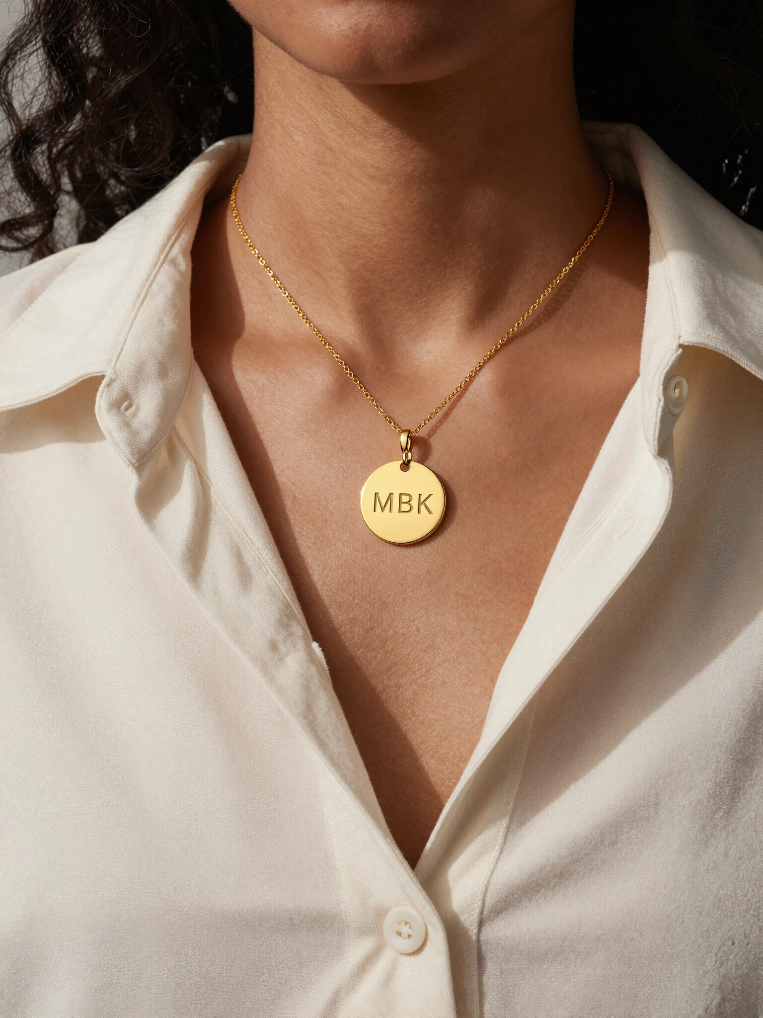 Woman wearing a gold circle necklace with engraved initials MBK, styled in a cream-colored shirt.